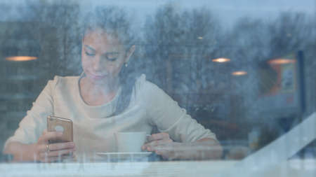 Smiling pretty woman with a cup of tea texting on smartphone in a cafeの写真素材