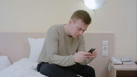 Worried young man typing an sms message on his cell phone sitting on bed at homeの写真素材