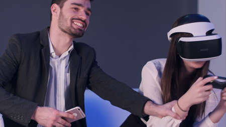 Smiling man with phone trying to distract his female partner from playing virtual reality gameの写真素材