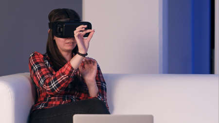 Laughing girl sitting on the sofa and using laptop via virtual reality glassesの写真素材