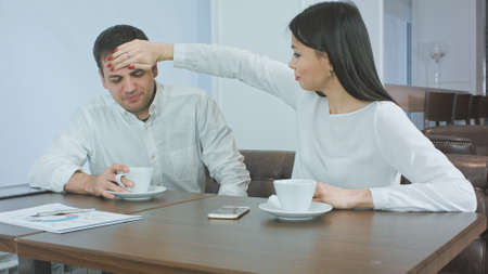 Sick businessman sneezing while worried female partner checking his head temperatureの写真素材