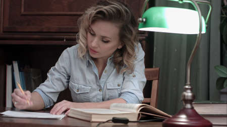 Young woman with a book sits down to the table, turns the green lamp on and starts workingの写真素材