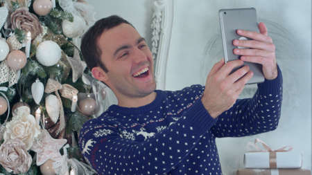 Young handsome man in a Christmas knitted sweater taking selfies on a tabletの写真素材