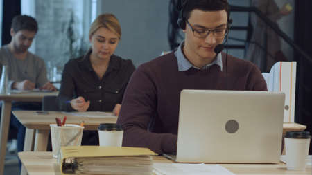 Happy cheerful young worker of call center in head-phones smiling and working on laptop
