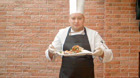 Friendly chef in uniform present a plate with seafood salad, talking to a cameraの写真素材
