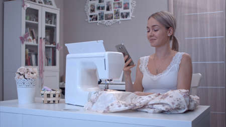 Young woman happily talking on the phone while sitting at the table next to sewing machineの写真素材