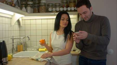 Happy couple talking and using smartphone while cooking in kitchen at homeの写真素材