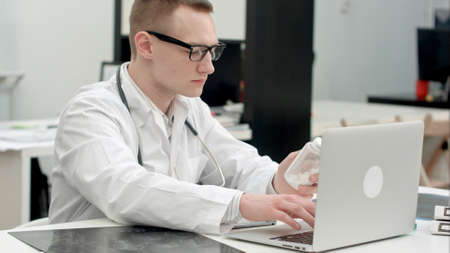 Male doctor holding jar of pills and typing prescription on laptopの写真素材