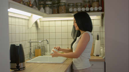 Young woman washing dirty dishes with soap and talking to someone in home kitchenの写真素材