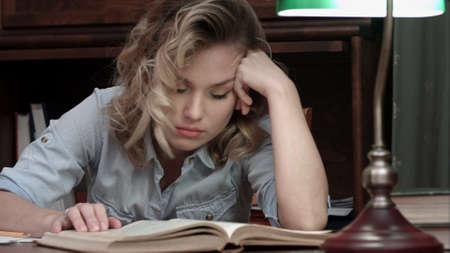 Tired young woman falling asleep over a book while sitting at the table after long day of workの写真素材