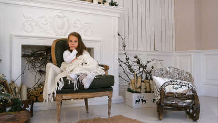 Little girl cozily wrapped in a blanket sitting on a chair near the Christmas treeの写真素材