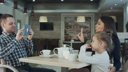 Happy father taking photo of his wife kissing their daughter by smartphone at restaurant.の写真素材