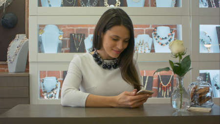 Nice female seller texting messages on smartphone and cute smiling in a jewelry storeの写真素材
