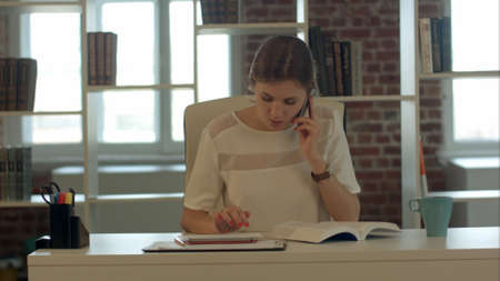Beautiful young blond woman talking on mobile phone and reading bookの写真素材