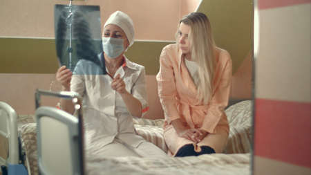 Professional doctor holding x-ray and talking to young female patient sitting on bed