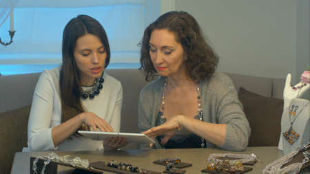 Young female seller and attractive woman select jewellery using tablet sitting on comfortable sofa in a jewelry storeの写真素材