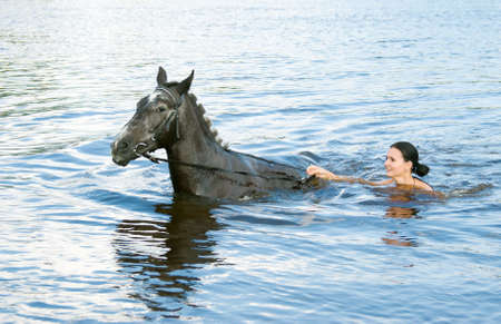 young woman swimming winth her black stallion in riverの写真素材