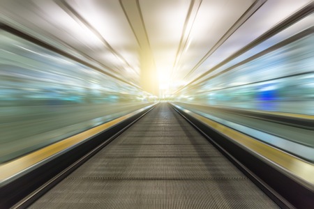 Perspective wide angle black and white view of modern light blue illuminated and spacious high-speed moving escalator with fast blurred trail of handrail in vanishing traffic motionの写真素材