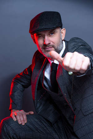 A man in a classic suit and coat looks directly into the camera and poses for a photographer. Shows...