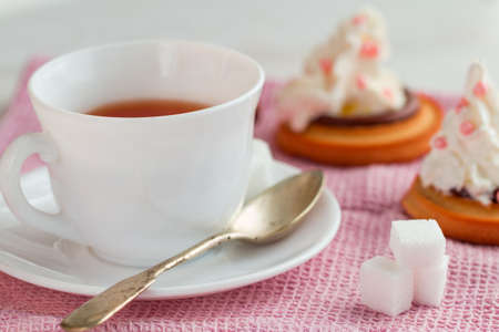 white china cup of black tea with teaspoon, cubic sugar and fresh biscuit cakes with whipped creamon pink kitchen towelの写真素材