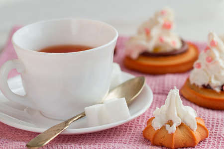 white china cup of black tea with teaspoon, cubic sugar and fresh biscuit cakes with whipped creamon pink kitchen towelの写真素材