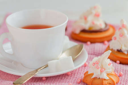 white china cup of black tea with teaspoon, cubic sugar and fresh biscuit cakes with whipped creamon pink kitchen towelの写真素材