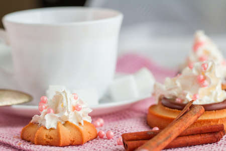 white china cup of black tea with teaspoon, cubic sugar and fresh biscuit cakes with whipped creamon pink kitchen towelの写真素材