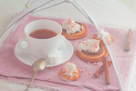 white china cup of black tea with teaspoon cubic sugar and fresh biscuit cakes with whipped cream on pink kitchen towel under net umbrella coverの写真素材