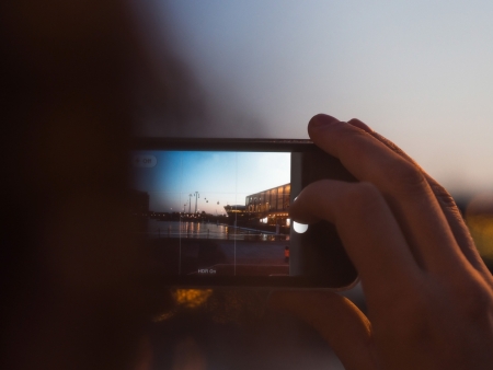 Over the shoulder view at dusk of a person holding the display of a digital device and photographing the sunsetの写真素材