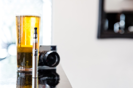 Full e-cigarette in a pub or bar standing on a reflective counter with a full pint of lager beer and a digital camera with copyspaceの写真素材