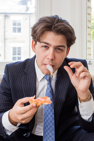 Smart attractive young businessman in a suit sitting in his urban office smoking while eating a snackの写真素材
