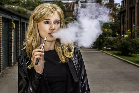 Pretty blond woman smoking an e-cigarette standing in a street in a leather jacket exhaling a cloud a smoke from her mouth while looking off to the sideの写真素材