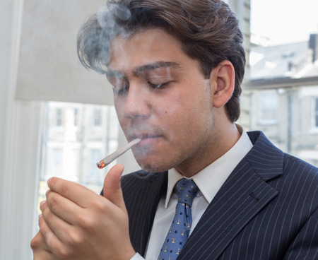 Young businessman lighting up a cigarette with a lighter in his cupped hands puffing smoke while standing in an urban office with windowsの写真素材