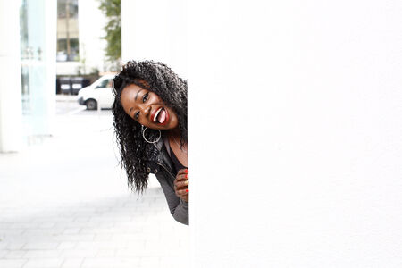 Playful young attractive African woman with a vivacious smile peeking around a pillar or column on an undercover urban walkwayの写真素材