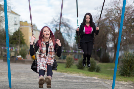 Front View of Two Young Funky Women Swinging on Swings and Chatting at Outdoor Playgroundの写真素材