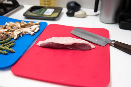Portion of raw meat with a sharp kitchen cleaver and fresh asparagus and mushrooms on chopping boards in the kitchen ready to prepare dinnerの写真素材