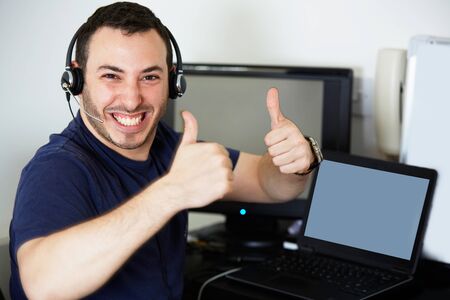 Happy call centre worker giving a thumbs up and looking to camera in front of two screens and...