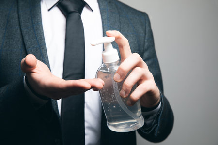 a man in a business suit and a mask cleans his hands with an antiseptic on a gray backgroundの写真素材