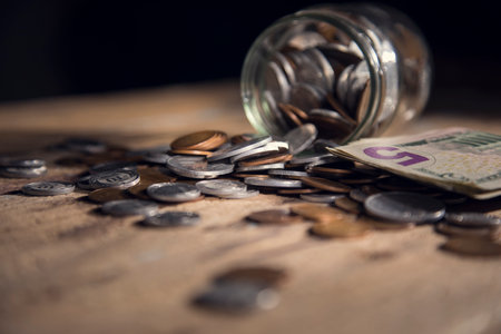 Coins are spilled from a glass bottle on a wooden tableの写真素材