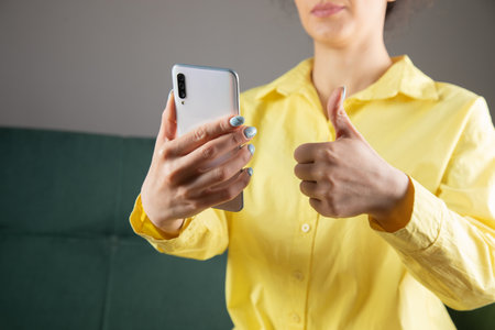 young woman holds her phone and shows her thumb up sitting on the couchの写真素材