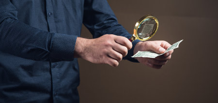 a man with a magnifying glass looks at money on a brown backgroundの写真素材