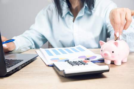 a woman sits in front of a work table and throws money into a piggy bank on a gray backgroundの写真素材
