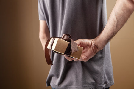 man hiding gift box from behind on brown background