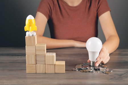 holds a light bulb near a coin and a rocket stands on a ladder of cubesの写真素材