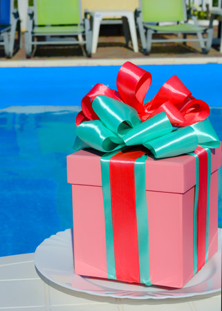Gift box for party decoration. The pink box is decorated with green and crimson ribbons on the background of the pool. Place for text.の写真素材