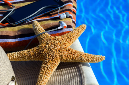 Women's hat, mobile phone and headphones next to the pool. Starfish with women's accessories.