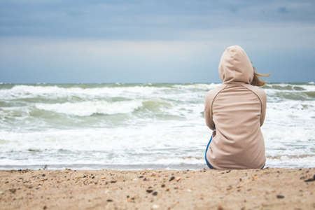 young woman sitting alone on the shore of the stormy sea in cold weather and finding reflects the...