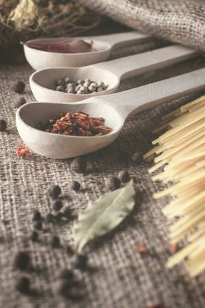 wooden spoons lie in a row with spices for cooking: red peppers, white and black pepper peas, garlic, bay leaf, dill and spaghettiの写真素材