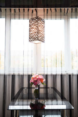A Bucket of peony flowers standing on the glass table under the curtains near the windowの写真素材