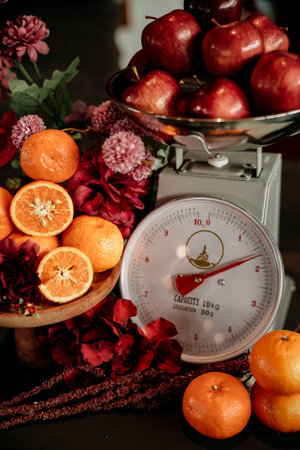 apples and oranges fruit on a scale with colorful flower decorations on a wooden tableの写真素材